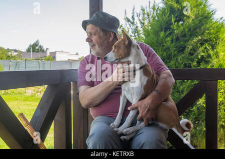 Happy senior man taking his lovely basenji dog and watching both while sitting on summer veranda Stock Photo