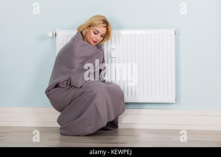 Young Woman With Blanket Suffering From Cold Sitting Near Heating Radiator At Home. Energy Saving Concept Stock Photo