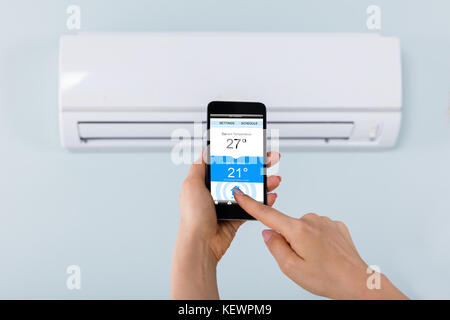 Close-up Of Person Hand Setting Temperature Of Air Conditioner Using Mobilephone At Home Stock Photo