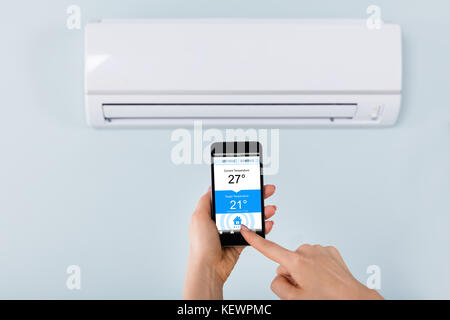 Close-up Of Person Hand Setting Temperature Of Air Conditioner Using Mobilephone At Home Stock Photo