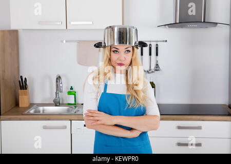 Portrait Of An Annoyed Young Woman With Cooking Pan On The Head Standing In Kitchen Stock Photo