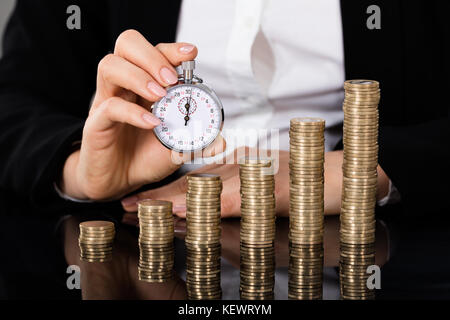Businesswoman Showing Stopwatch In Front Of Stacked Coin On Desk. Quick Money Concept Stock Photo