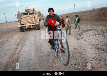 A young Afghan Boy pulls a wheelie as he rides in front of a British Army Mastiff armoured vehicle Stock Photo