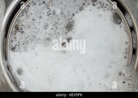 White foam with bubbles of cleane in a washbasin. Drain hole with soap bubbles in metal sink, macro view. Mechanically adjustable drain plug closeup. Stock Photo