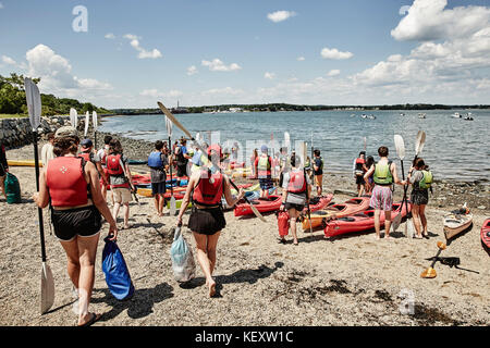 Group of people walking and standing with paddles beside kayaks lying on sea coast, Portland, Maine, USA Stock Photo