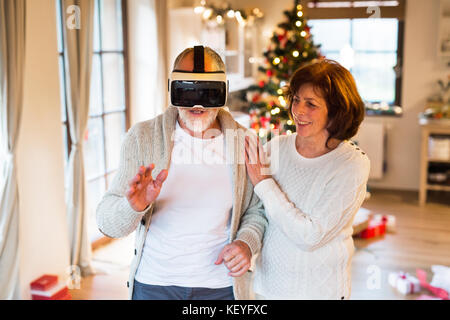 Senior couple in front of illuminated Christmas tree inside their house trying on VR glasses. Stock Photo