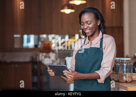 Smiling young African entrepreneur using a tablet in her cafe Stock Photo