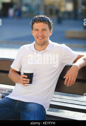 Portrait of a young dark-haired, curly-haired man. The guy is sitting alone on a bench with a paper cup of coffee in a hand. He smiling to his thought Stock Photo