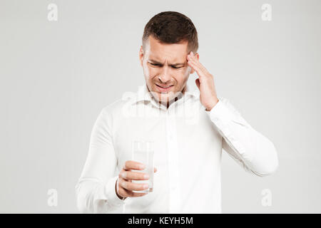 Photo of sad young man with headache dressed in white shirt isolated over grey wall background. Looking aside holding glass full of water. Stock Photo