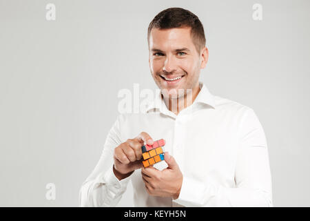 Image of smiling young man dressed in white shirt isolated over grey wall background. Looking camera solve Rubik's cube. Stock Photo