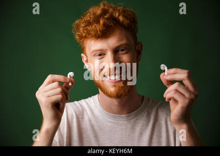 Close-up of young smiling curly redhead bearded young man in white tshirt, showing airpods, over green background Stock Photo