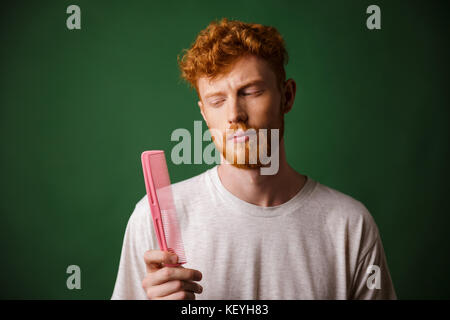 Young readhead beardy man looking at pink comb, over green background Stock Photo