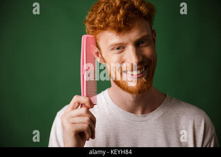 Smiling young readhead beardy man showing pink comb, over green background Stock Photo