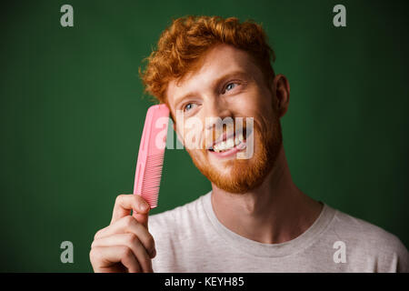 Close-up portrait of smiling young readhead beardy man with pink comb, over green background Stock Photo