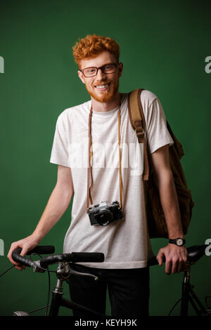 Young happy readhead bearded man with backpack and retro camera, standing on bicycle isolated over green background Stock Photo