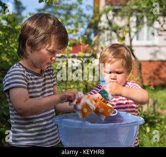 Two small boys help to wash dishes outdoors in the village, in the country. Focus on the cup Stock Photo