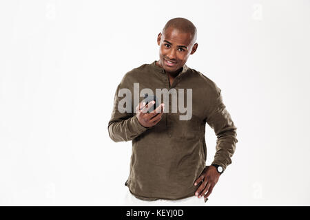 Smiling african man holding arm on hip and using smartphone while looking at the camera over white background Stock Photo