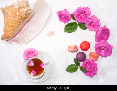 Flat-lay of a cup of tea with rose petals, pink sea shells, macarons and pink roses Stock Photo