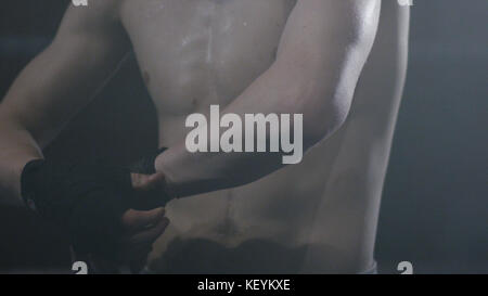 Close up of a boxer putting on straps preparing for combat on a dark background. Closeup male hand of boxer with red boxing bandages. Fists of fighter before the fight or training in sport gym. Stock Photo