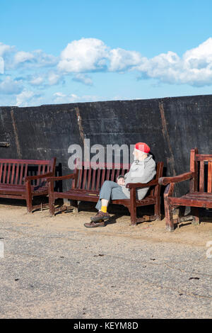 An elderly gentleman with a red woollen hat, sits on benches on the harbour arm at Broadstairs, Kent, in the shelter and afternoon autumn sun. Stock Photo