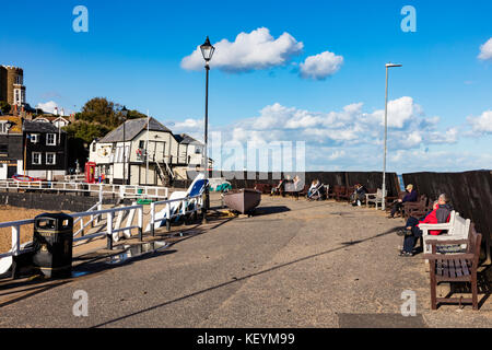 Groups of people, visitors and families sit on Broadstairs Harbour Arm and enjoy the Autumn sunshine in the shelter of a wall, Kent, UK Stock Photo