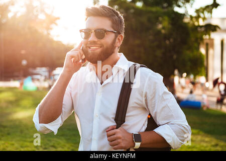 Happy bearded man in sunglasses standing outdoors while holding backpack and talking by smartphone Stock Photo