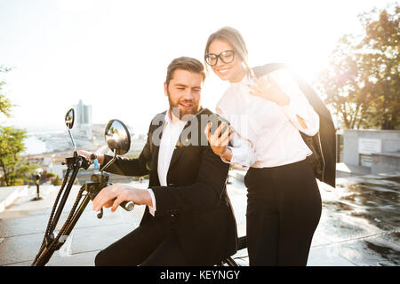 Smiling business woman standing near man in suit which sitting on modern motorbike outdoors while showing something on her smartphone Stock Photo