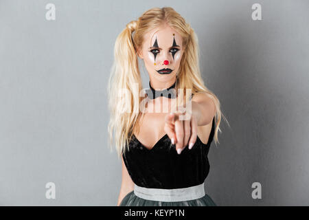 Portrait of a serious young blonde woman in halloween clown make-up posing while standing and pointing at camera isolated over gray background Stock Photo