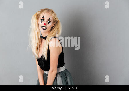 Portrait of a pretty blonde woman in halloween clown make-up posing while standing and looking away isolated over gray background Stock Photo