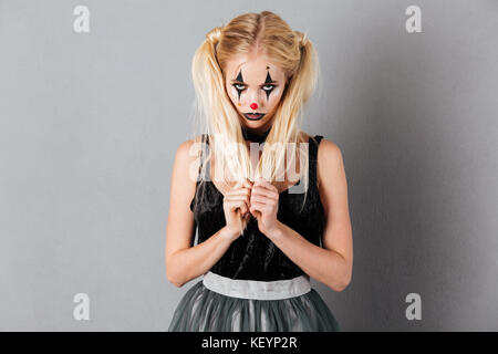 Portrait of a pretty blonde woman in halloween clown make-up posing while standing and looking at camera isolated over gray background Stock Photo