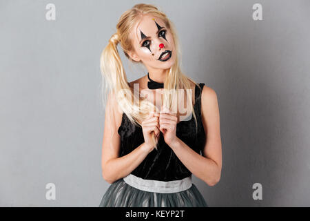 Portrait of a playful blonde woman in halloween clown make-up posing while standing and looking at camera isolated over gray background Stock Photo