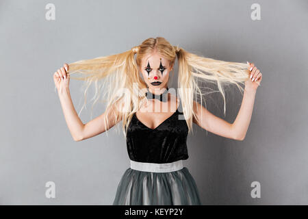 Portrait of a serious blonde woman in halloween clown make-up playing with hair while standing and looking at camera isolated over gray background Stock Photo