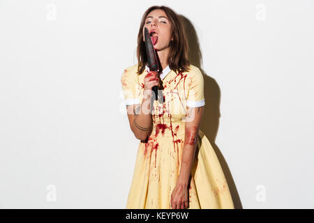 Pretty zombie woman in dress licking knife and looking at the camera over white background Stock Photo