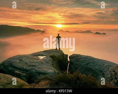 Man stands alone on the peak of rock. Hiker watching to autumn  Sun at horizon . Beautiful moment the miracle of nature. Colorful mist in valley. Man  Stock Photo