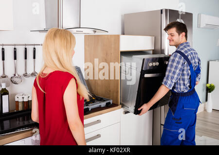 Male Worker Installing Oven For Repairing While Young Woman Standing In Kitchen Stock Photo
