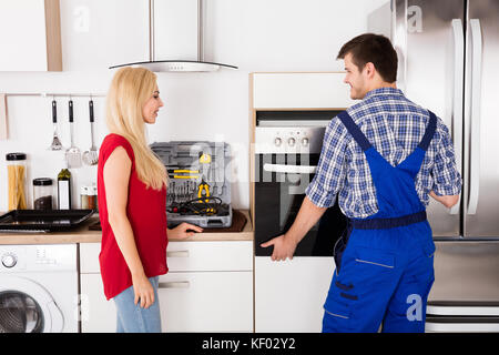 Male Worker Installing Oven For Repairing While Young Woman Standing In Kitchen Stock Photo