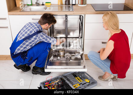 Young Woman Looking At Repairman Repairing Dishwasher In Kitchen Stock Photo
