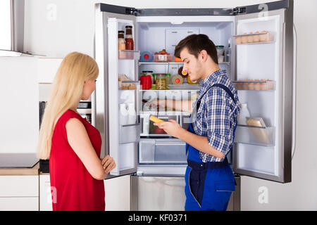 Young Woman Looking At Male Technician Checking Fridge With Digital Multimeter Stock Photo