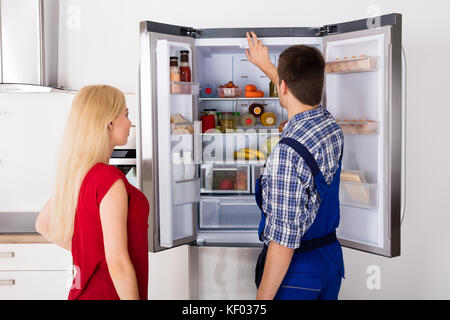 Woman Looking At Male Worker Repairing Refrigerator In Kitchen Room Stock Photo