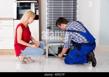 Woman Looking At Male Repairman Checking Fridge With Digital Multimeter At Home Stock Photo