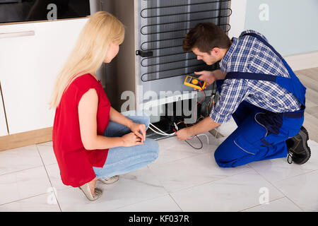 Woman Looking At Male Repairman Checking Fridge With Digital Multimeter At Home Stock Photo