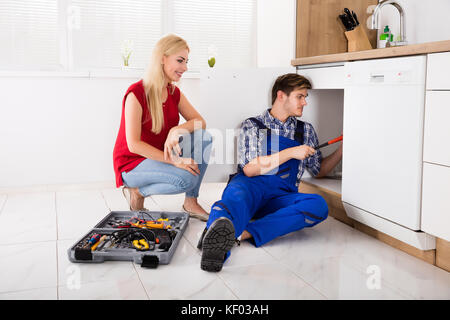 Young Woman Looking At Male Plumber Fixing Kitchen Sink Using Toolbox Stock Photo