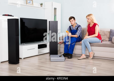 Male Technician Checking Home Theater TV Speaker In Front Of Woman On Couch At Home Stock Photo