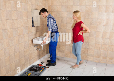 Young Handyman Installing Sink With Young Woman Standing In Bathroom Stock Photo
