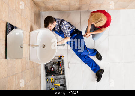 Woman Looking At Young Male Plumber Lying On Floor Fixing Sink In Bathroom Stock Photo