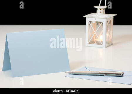 Still life with envelope, paper card and pen in front of burning candle in lamp on white board table and black background with blurred background Stock Photo