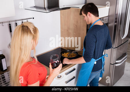 Young Woman Drinking Wine Looking At Her Husband Preparing Dinner In Kitchen Stock Photo