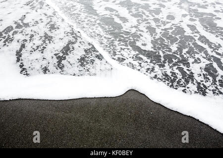 The sea spills foam over black sand as the tide comes in to a small beach. Stock Photo