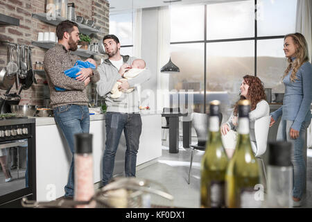Two wives watching husbands holding their babies in kitchen Stock Photo