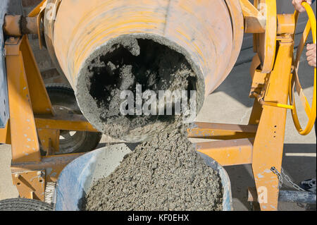 A close up image of a mans hands pouring wet cement into wheelbarrow from an electric mixer machine. Stock Photo